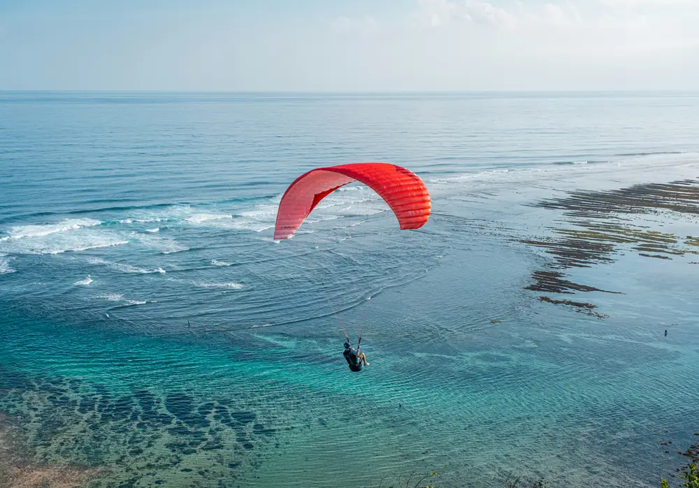 Paraglider flying over clear turquoise ocean waters along the Bali coast with Bali Assist Co