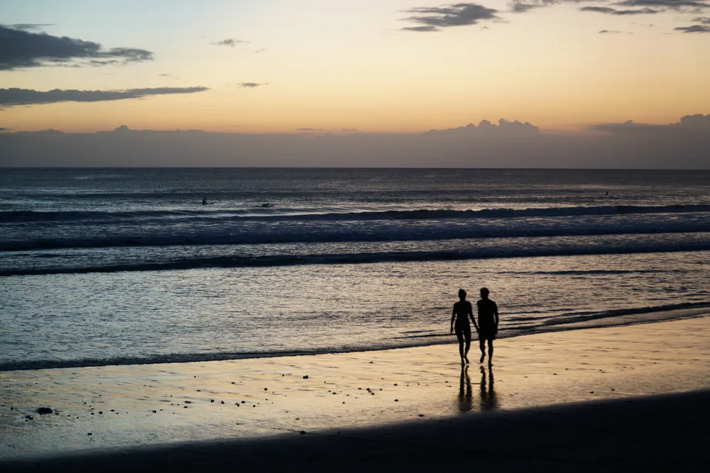 Couple standing by the ocean coast at golden hour sunset for The Discovery Bali tour package by Bali Assist Co
