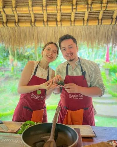 Couple wearing aprons holding freshly prepared Balinese dishes in a cooking class for The Discovery by Bali Assist Co