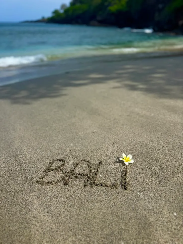BALI text written on sandy beach with a frangipani flower for Day 7 departure itinerary by Bali Assist Co