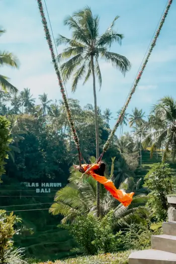 Tourist riding high swing with orange dress flowing over Ubud coconut trees for The Signature Journey by Bali Assist Co