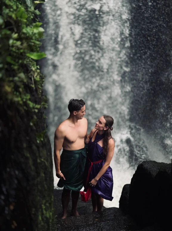Couple wearing traditional sarongs standing together in front of waterfall for private Melukat water purification ceremony in Bali by Bali Assist Co